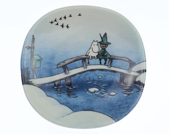 Op de afbeelding: Een wit keramiek bord met een blauwe en witte illustratie van twee personages op een brug over een bevroren meer. De personages zijn een wit wezen met een lange snuit en een groen wezen met een hoed. Er vliegen vogels in de lucht boven de brug.