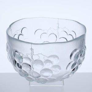 May include: A clear glass bowl with a textured surface and a design of raised, circular shapes resembling grapes. The bowl has a square shape with rounded edges and sits on a clear stand.