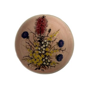 Könnte beinhalten: Ein runder, rosa Keramikteller mit handgemaltem Blumendesign. Die Anordnung zeigt eine zentrale rote, stachelige Blume, Gruppen gelber und weißer Blüten und blaue kugelförmige Blüten. Die Initialen "J.F." sind sichtbar.