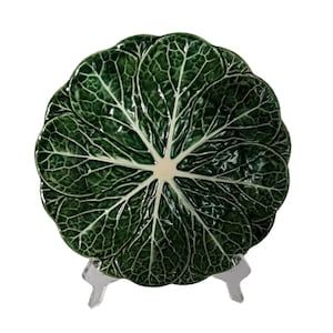 May include: A green ceramic serving dish shaped like a cabbage with detailed leaf veins. The dish is on a clear plastic stand.