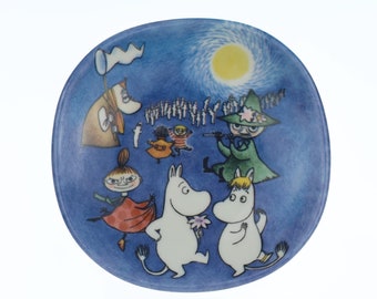 Op de afbeelding: Een blauw keramiek bord met een fantasievolle illustratie van Moomin-personages. De scène toont Moomin en zijn vrienden dansen onder een sterrenhemel met een gele maan. Het bord heeft een witte rand en een blauwe achtergrond.
