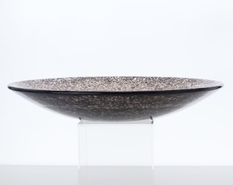 May include: A decorative, wide, shallow bowl with a speckled pattern in shades of brown and gray. The bowl has a dark rim and is displayed on a clear, square stand. The bowl's design is modern and elegant.