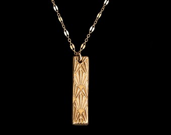 May include: A gold necklace with a geometric patterned pendant. The pendant is a rectangular shape with a repeating pattern of sunbursts.