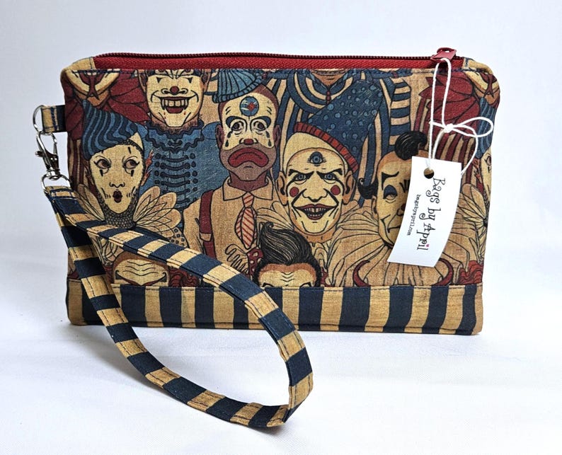 May include: A rectangular clutch with a zipper closure and a wrist strap. The clutch features a print of vintage circus clowns in shades of red, blue, and tan. The wrist strap has a navy blue and tan striped pattern. A tag reads "Bags by Appell".