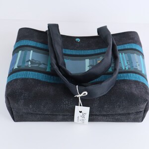 May include: A dark gray tote bag with a textured pattern. The bag features a forest scene design in shades of blue and teal across the top, with matching teal stripes. It has dark gray handles and a small tag.