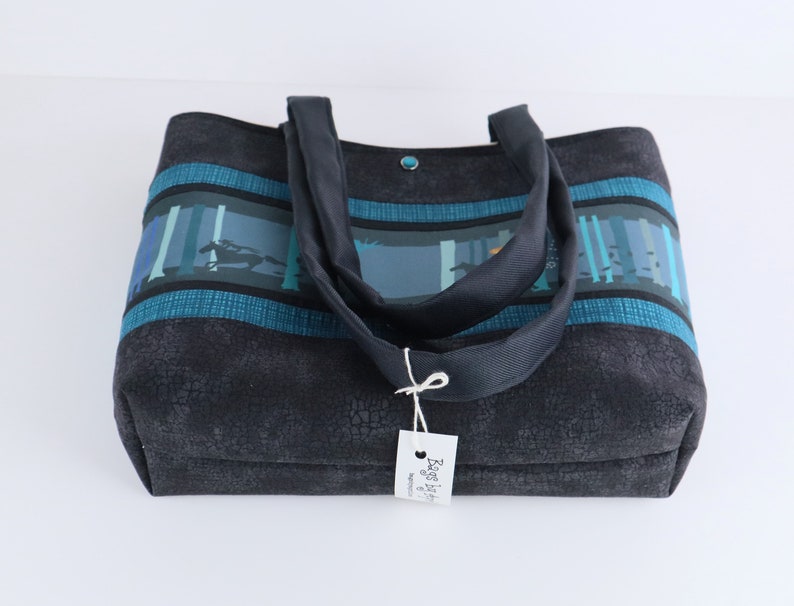May include: A dark gray tote bag with a textured pattern. The bag features a forest scene design in shades of blue and teal across the top, with matching teal stripes. It has dark gray handles and a small tag.