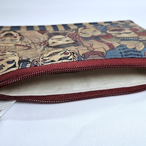 May include: A rectangular pouch with a burgundy zipper and a beige interior. The exterior features a repeating pattern of stylized faces in shades of tan, blue, and red. The pouch is open, revealing the interior lining.