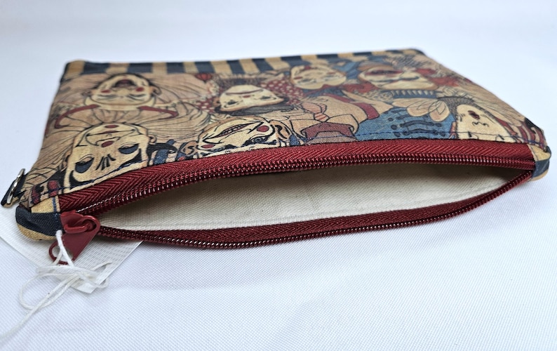 May include: A rectangular pouch with a burgundy zipper and a beige interior. The exterior features a repeating pattern of stylized faces in shades of tan, blue, and red. The pouch is open, revealing the interior lining.