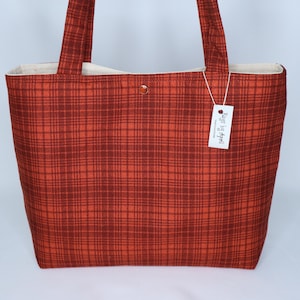 May include: A red and brown plaid tote bag with matching handles. The bag has a light-colored interior and a small tag attached. The bag is made of fabric and has a button closure.