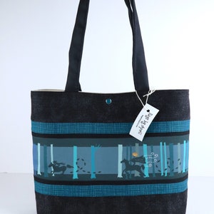 May include: Black tote bag with a forest scene design. The bag features a black background with teal and blue accents. The design includes a person riding a horse through a forest. The bag has black handles and a button closure.