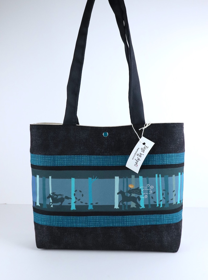 May include: Black tote bag with a forest scene design. The bag features a black background with teal and blue accents. The design includes a person riding a horse through a forest. The bag has black handles and a button closure.