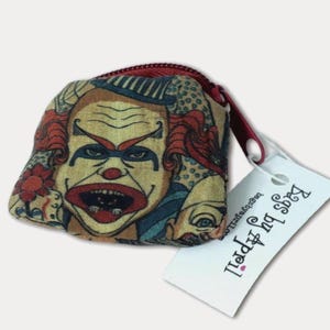 May include: A small, square coin purse with a zipper closure. The purse features a vintage-style illustration of a clown's face in shades of yellow, red, and blue. A white tag with the text "Bags by Appell" is attached.