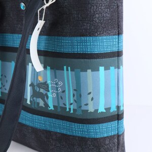 May include: A tote bag with a forest scene design. The bag features a black textured exterior with teal and blue stripes. The central panel depicts a forest with trees and a silhouette of a horse and rider. A tag hangs from the handle.