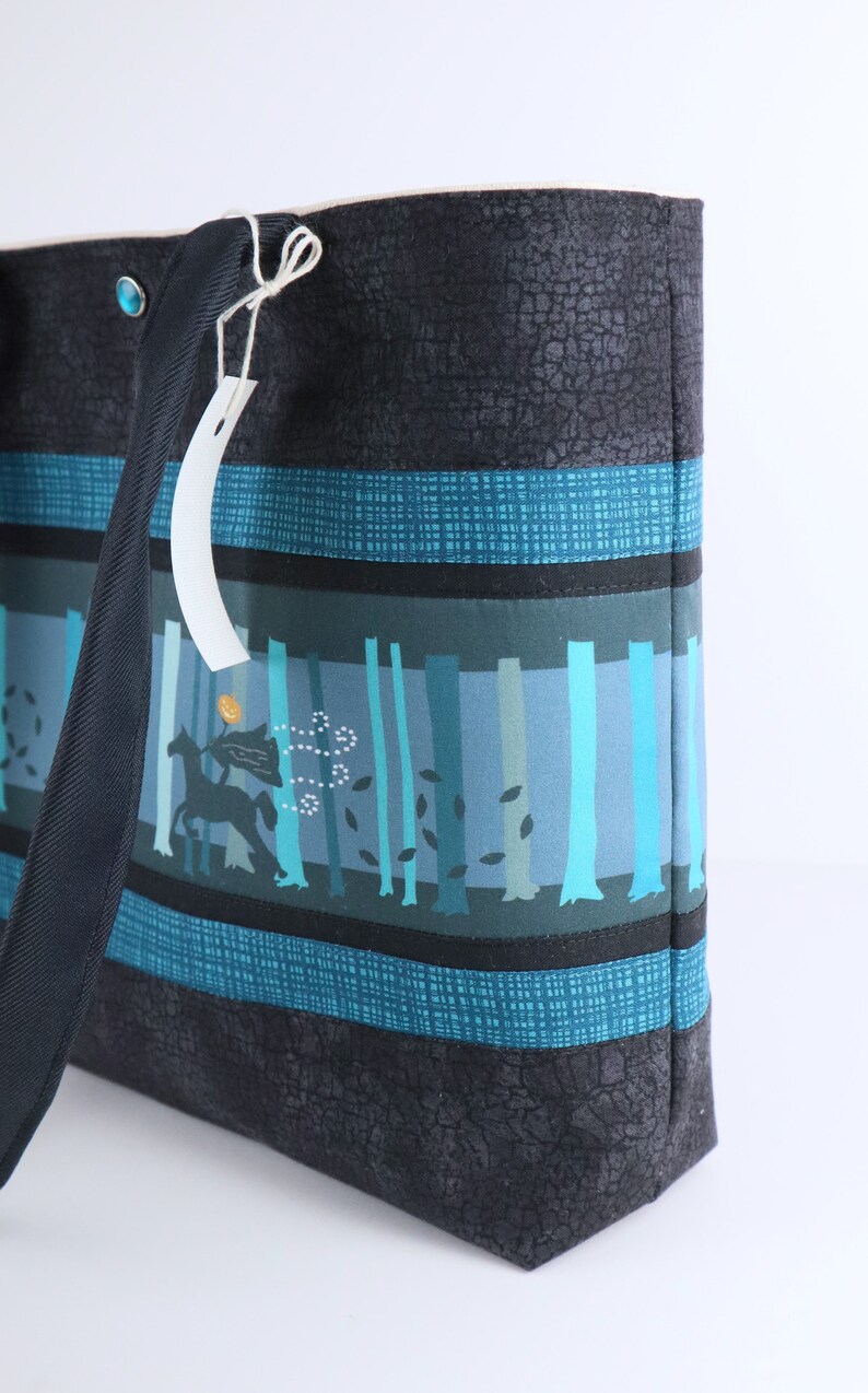 May include: A tote bag with a forest scene design. The bag features a black textured exterior with teal and blue stripes. The central panel depicts a forest with trees and a silhouette of a horse and rider. A tag hangs from the handle.