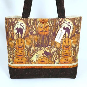 May include: A Halloween-themed tote bag with a repeating pattern of jack-o'-lanterns, black cats, and ghosts in a forest setting. The bag has brown handles and a dark brown base. A tag with text is attached to the bag.