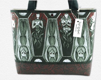 May include: A tote bag featuring a repeating pattern of coffins containing bats and figures with crossed arms, set against a dark red and black background. The bottom panel displays a dark red rose print. A white tag reads Bags by April.