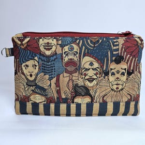 May include: A rectangular zippered pouch featuring a collage of vintage clown faces in shades of red, blue, and beige. The pouch has a red zipper and a small metal loop on the side. The bottom of the pouch has blue and beige stripes.