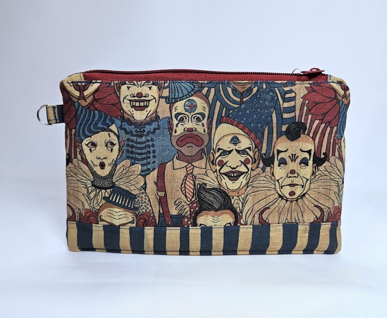 May include: A rectangular zippered pouch featuring a collage of vintage clown faces in shades of red, blue, and beige. The pouch has a red zipper and a small metal loop on the side. The bottom of the pouch has blue and beige stripes.