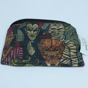 May include: A zippered pouch featuring a multi-colored graphic print of classic horror movie monsters including a vampire, a mummy, and a werewolf. The fabric has a dark, muted color palette and a black zipper closure along the top edge.