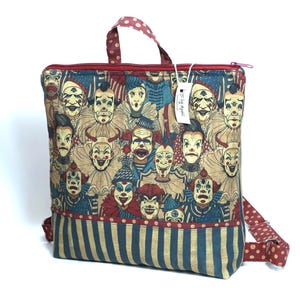 May include: A square fabric backpack featuring a repeating pattern of various clown faces in shades of red, blue, and beige. The bag has a red zipper closure, a top handle with red and white polka dots, and a bottom panel with vertical blue stripes.