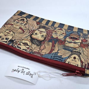May include: A rectangular, zippered pouch featuring a repeating pattern of stylized clown faces in shades of tan, red, and blue. The pouch has a dark red zipper and a tag that reads "Bags by Appell".