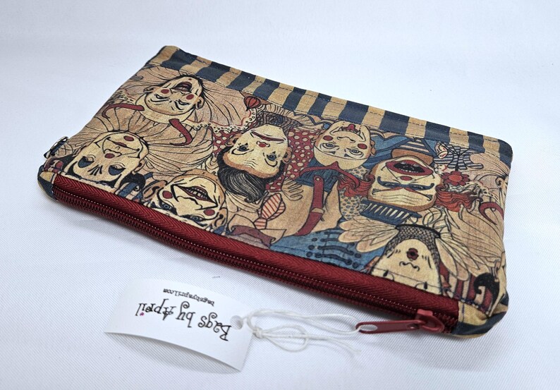 May include: A rectangular, zippered pouch featuring a repeating pattern of stylized clown faces in shades of tan, red, and blue. The pouch has a dark red zipper and a tag that reads "Bags by Appell".