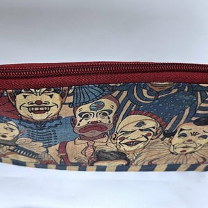 May include: A rectangular, zippered pouch featuring a repeating pattern of vintage-style clown faces in red, blue, and beige. The zipper and trim are a deep burgundy color. A small loop is attached to one end.