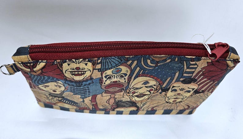 May include: A rectangular, zippered pouch featuring a repeating pattern of vintage-style clown faces in red, blue, and beige. The zipper and trim are a deep burgundy color. A small loop is attached to one end.
