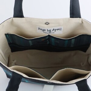 May include: Open tote bag with a dark blue exterior and cream-colored interior. The bag features multiple interior pockets with a teal and black plaid pattern. The handles are dark blue, and the bag has a magnetic snap closure. The label inside reads "Bags by April".