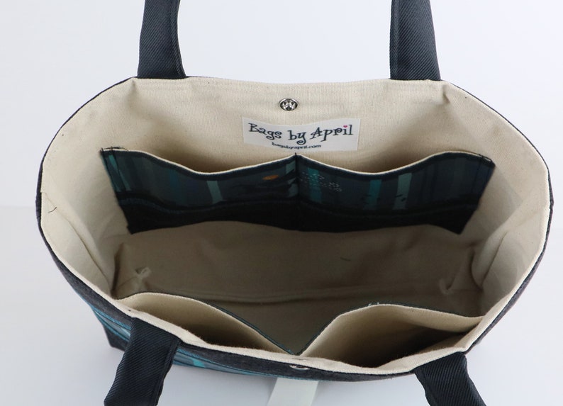 May include: Open tote bag with a dark blue exterior and cream-colored interior. The bag features multiple interior pockets with a teal and black plaid pattern. The handles are dark blue, and the bag has a magnetic snap closure. The label inside reads "Bags by April".