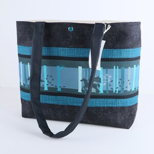 May include: A tote bag with a forest scene design. The bag features a dark blue textured fabric with teal and blue stripes. The center panel depicts a forest with trees and a running horse. The bag has a snap closure and dark blue handles.