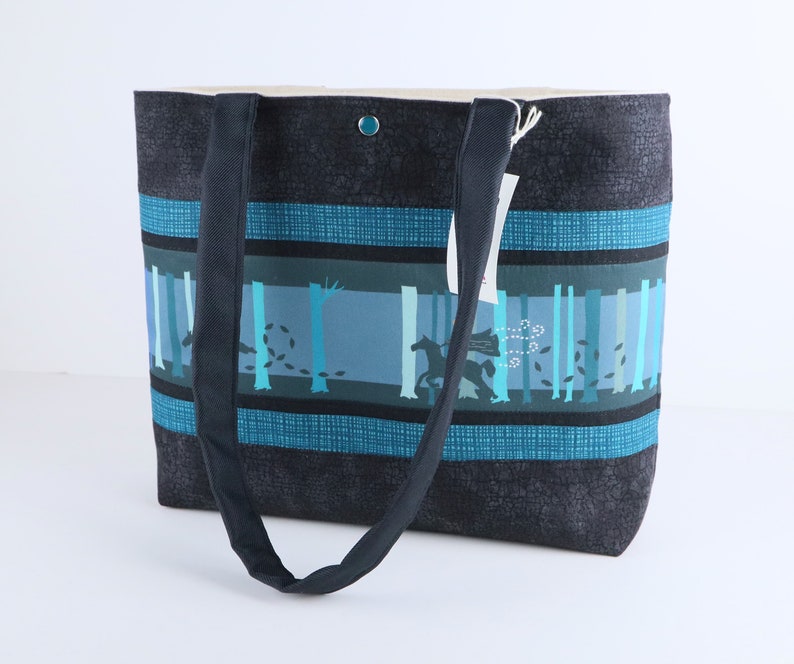 May include: A tote bag with a forest scene design. The bag features a dark blue textured fabric with teal and blue stripes. The center panel depicts a forest with trees and a running horse. The bag has a snap closure and dark blue handles.