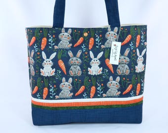 May include: A blue tote bag with a white and orange bunny and carrot pattern. The bag has a white, orange, and green striped trim and a blue denim bottom. The bag has two blue handles and a tag that says "Handmade by April".