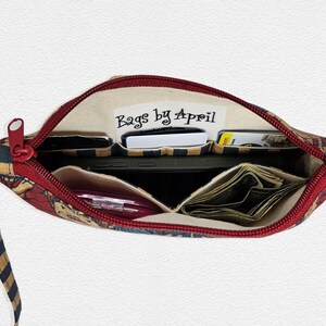 May include: Open zippered pouch with a red zipper and wrist strap. The interior reveals multiple compartments holding a smartphone, cash, cards, and a pink pen. The pouch is patterned with a mix of colors and the text "Bags by April".