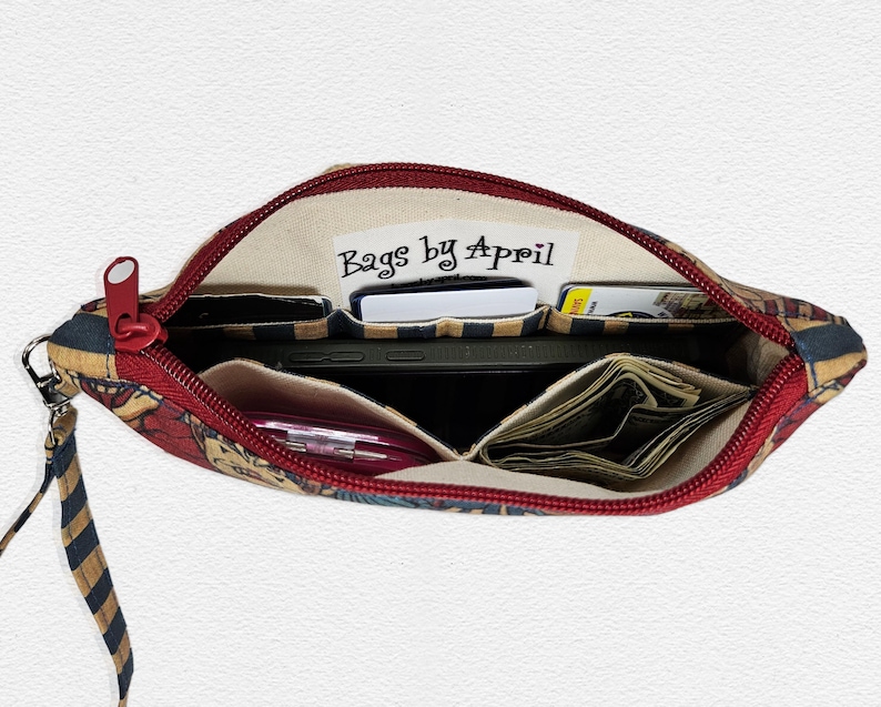 May include: Open zippered pouch with a red zipper and wrist strap. The interior reveals multiple compartments holding a smartphone, cash, cards, and a pink pen. The pouch is patterned with a mix of colors and the text "Bags by April".