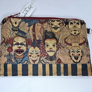 May include: A rectangular zippered pouch featuring a repeating pattern of vintage-style clown faces in shades of beige, red, and blue. The bottom of the pouch has a striped pattern in navy and tan. The zipper is red, and the tag reads "Lags by Aspen".