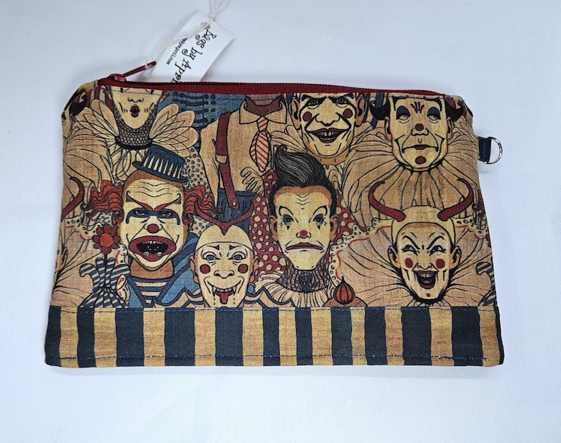 May include: A rectangular zippered pouch featuring a repeating pattern of vintage-style clown faces in shades of beige, red, and blue. The bottom of the pouch has a striped pattern in navy and tan. The zipper is red, and the tag reads "Lags by Aspen".