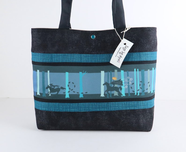 May include: A tote bag with a dark blue textured exterior and a forest scene design. The forest scene features silhouettes of riders on horseback and trees in shades of blue and teal. The bag has dark blue handles and a button closure.