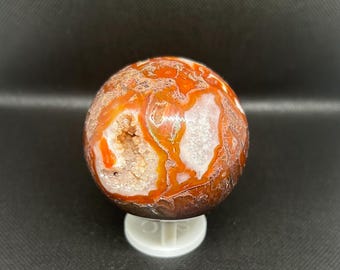 May include: A polished orange and white agate sphere with a natural crystal formation. The sphere is displayed on a white stand.