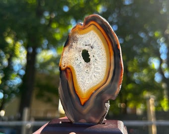 May include: A polished agate slice featuring concentric bands of dark gray, amber, and white, with a central crystalline geode cavity. The stone is mounted vertically on a rectangular dark brown wooden base, displaying natural mineral layers.