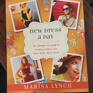 Op de afbeelding: Boekomslag met de titel "new dress a day" en de ondertitel "The Ultimate DIY Guide to Creating Fashion Dos from Thrift-Store Don'ts". De auteur is Marisa Lynch. De omslag toont vier foto's van vrouwen die verschillende outfits dragen.