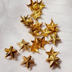 May include: A collection of gold-colored metal star-shaped embellishments. The stars are scattered on a white fabric surface.