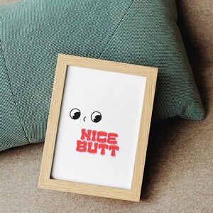 May include: A framed print with a cartoon design. The print features two black eyes and the red text "NICE BUTT" on a white background. The frame is made of light-colored wood. A teal pillow is in the background.