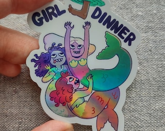 May include: A die-cut sticker featuring three colorful mermaids with sharp teeth and expressive faces. The text GIRL DINNER is printed in dark blue above the mermaids, with a single human foot dangling from a green water ripple graphic.