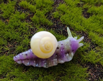 May include: A small decorative snail figurine featuring a translucent purple body with dark purple spots and a smooth, cream-colored spiral shell. The snail has two distinct tentacles extending from its head and a textured, segmented underside.