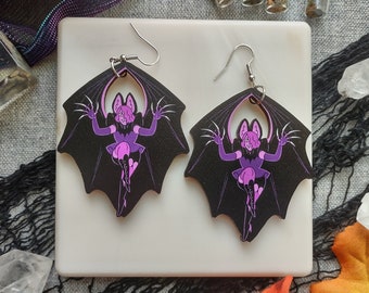 May include: A pair of black bat-shaped earrings with a purple cartoon character design. The earrings have silver hooks and are displayed on a white square surface. The background includes crystals, leaves, and small glass bottles.