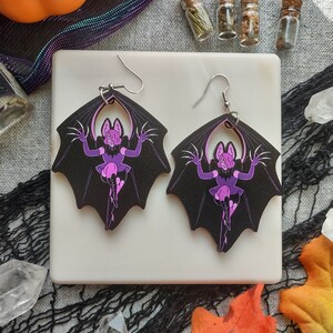 May include: A pair of black bat-shaped earrings with a purple cartoon character design. The earrings have silver hooks and are displayed on a white square surface. The background includes crystals, leaves, and small glass bottles.