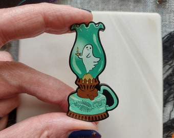 May include: A teal and brown decorative pin in the shape of an antique oil lamp. The lamp features a cartoon ghost holding a candle inside the glass. The pin has a black outline and is held by a person.