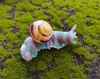 May include: A decorative snail figurine featuring a translucent, glittery body with swirling pastel pink and teal colors. The snail has a realistic, polished brown and cream-colored spiral shell mounted on its back, with two distinct eye stalks.