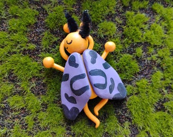 May include: A wooden insect figurine with a yellow body, round head, and black fuzzy antennae. The insect features two large, flat, light purple wings decorated with irregular black spots. It has thin yellow limbs and a painted face with closed eyes.